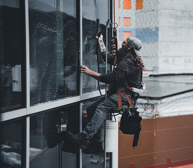 A window cleaner suspended from a harness, scrubbing a large glass pane, with a construction site visible in the background.