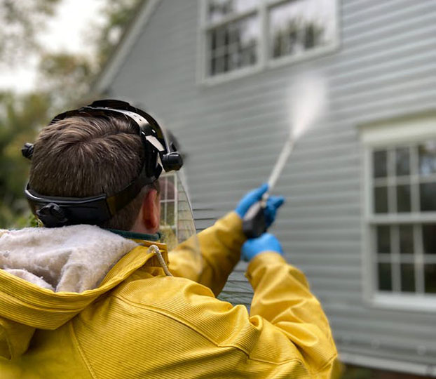 A person in a yellow raincoat and protective gear sprays a house's siding with a cleaning solution from a nozzle.